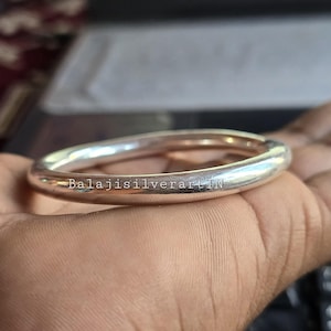 May include: A silver bangle bracelet with a smooth, rounded design. The bracelet is held in a hand and features the text "BalajisilverartIN". The bangle is a bright, reflective silver color.