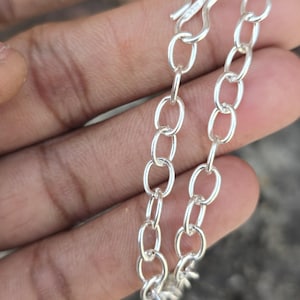 Puede incluir: Primer plano de un collar de cadena plateada con eslabones ovalados. La cadena está hecha de un metal brillante y reflectante y se muestra sobre un fondo neutro. El collar es una joya sencilla y elegante.
