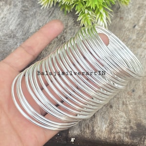 May include: A collection of silver bangles, held in a hand, with the text "Balajisilverart IN" visible. The bangles are circular and polished, reflecting light. The background is a wooden surface with green leaves.