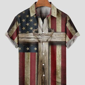 May include: Short-sleeved button-down shirt featuring a design that combines the American flag with a wooden cross and a crown of thorns. The shirt has a collared neckline and a button-up front. The design incorporates red, white, and blue colors.