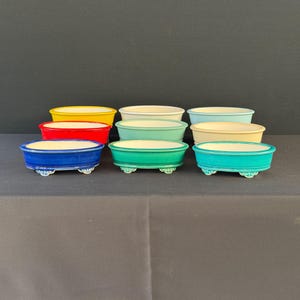 May include: A collection of nine oval ceramic bonsai pots arranged in three rows. The pots feature a variety of solid colors including yellow, red, dark blue, white, light blue, teal, cream, and green, each with small decorative feet.