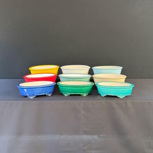 May include: A collection of nine oval ceramic bonsai pots arranged in three rows. The pots feature a glossy finish in various solid colors including yellow, white, light blue, red, mint green, beige, dark blue, forest green, and turquoise.