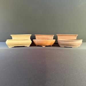 May include: A collection of six bonsai pots in various shapes and sizes. The pots are made of terracotta and have a natural, earthy color. Some pots have a rectangular shape, while others are oval or round. The pots are displayed on a dark surface.