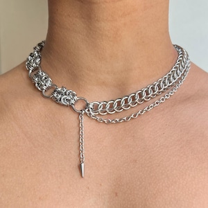 May include: A silver-toned chainmail necklace with a layered design. The necklace features a series of interconnected rings and a dangling chain with a cone-shaped pendant. The jewelry is designed to be worn around the neck.