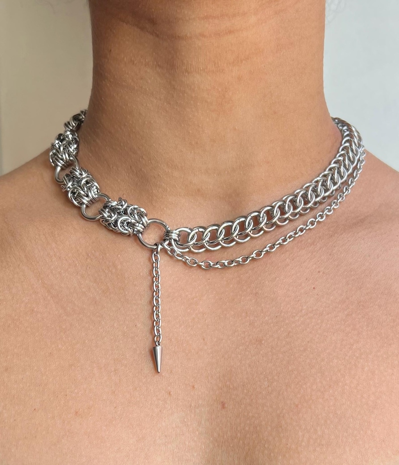 May include: A silver-toned chainmail necklace with a layered design. The necklace features a series of interconnected rings and a dangling chain with a cone-shaped pendant. The jewelry is designed to be worn around the neck.
