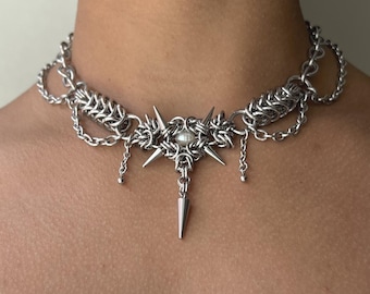 May include: A silver-toned chainmail choker necklace featuring intricate metal links, a central star-shaped pendant with a small white pearl at its core, and several sharp, pointed metal spikes extending from the center and the bottom.