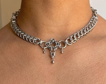 May include: A silver-toned chainmail choker necklace featuring a series of interlocking metal rings. The center of the necklace displays a decorative geometric pendant element with three small dangling metal spheres attached to the bottom edge.