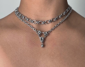 May include: A double-layered silver-toned metal chain necklace. The top layer features oval links with small crossbars, while the bottom layer consists of a standard link chain connected to a central ring with a small dangling metal sphere charm.