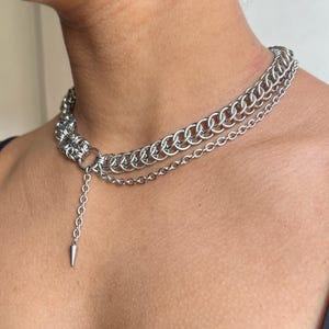 May include: A silver-toned chainmail choker necklace with a layered design. The necklace features multiple rows of interlocking rings and a dangling chain with a pointed cone pendant. The jewelry is worn around the neck.