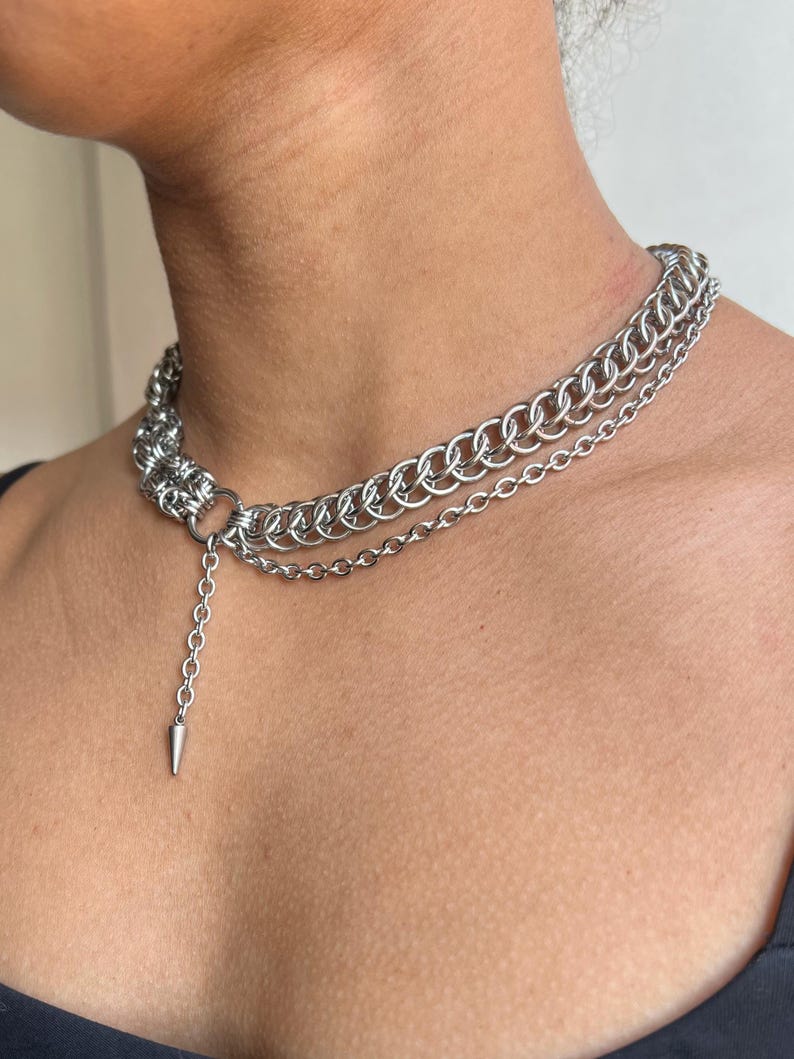 May include: A silver-toned chainmail choker necklace with a layered design. The necklace features multiple rows of interlocking rings and a dangling chain with a pointed cone pendant. The jewelry is worn around the neck.