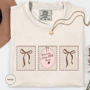 May include: Ivory-colored t-shirt with a crew neck and the Comfort Colors brand tag. The shirt features three decorative panels with brown bows and the text "brewing a tiny little bean" in a pink frame. A gold star pendant necklace is draped across the shirt.