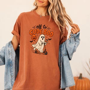 May include: A rust-colored t-shirt featuring a graphic of a ghost riding a scooter while holding a coffee cup and a jack-o'-lantern balloon. The text above the ghost reads "off to CODE BOO" in orange and black lettering, accompanied by small bats.
