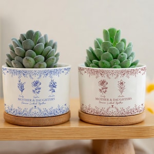 May include: Two white ceramic planters with succulents, each featuring floral designs and the text "Mother & Daughters Forever Linked Together." One planter has blue accents, the other burgundy. Both sit on wooden saucers.