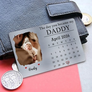 May include: A silver metal wallet card with a photo of a baby and the text "The day you became my DADDY April 2026." The card includes a calendar with the date April 30 highlighted with a heart, and the name "Andy".