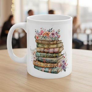 May include: White ceramic mug featuring a stack of books with titles like "Fantasy," "Body Love," and "Thriller." The books are adorned with colorful floral arrangements. The mug has a white handle and is set on a wooden surface.