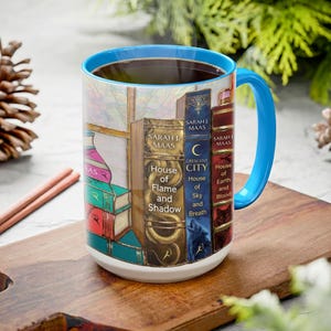 May include: A white ceramic mug with a blue handle and rim, filled with dark liquid. The mug features a design of stacked books with titles by Sarah J. Maas. The books are in various colors, including red, blue, and gold. The mug sits on a wooden surface.