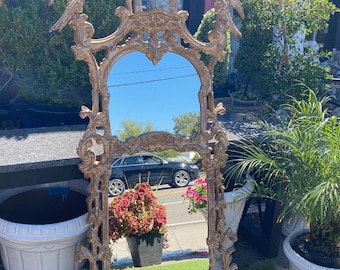 May include: A large, ornate gold-colored mirror with a detailed, carved frame. The mirror has a rectangular shape with a curved top and bottom. Two bird sculptures adorn the top corners. The mirror is standing on a green surface.