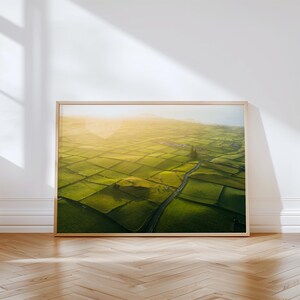 May include: A framed landscape print featuring a vibrant aerial view of green fields divided into geometric shapes. A road winds through the fields, and the sun shines brightly in the upper portion of the image, creating a warm, inviting atmosphere.