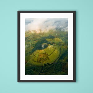 Peut inclure: Photographie aérienne encadrée d'un cratère volcanique et de terres agricoles environnantes. L'image présente un paysage vert luxuriant avec un cratère central circulaire et un réseau de champs. Le ciel est partiellement voilé de nuages blancs.
