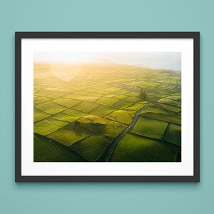 May include: Framed aerial photograph of a patchwork of green fields, divided by stone walls, under a bright, golden sky. A winding road cuts through the landscape. The artwork is displayed on a teal wall.