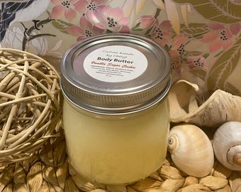 May include: A jar of body butter with a silver lid and a label that reads "Vanilla Sugar Cookie." The jar is surrounded by decorative elements, including seashells and a woven ball. The body butter appears to be a light yellow color.