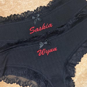 May include: Two black underwear with red embroidered names, Saskia and Wynn. The underwear have a ribbed texture, black lace trim, and small black bows. The background is a light brown.