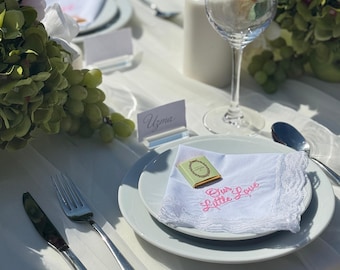 Pode incluir: Uma mesa de casamento com toalha branca, pratos e talheres. Uma toalha de mesa branca com bordado rosa com a frase "Our Little Love" está sobre um prato. Uma pequena caixa verde e dourada está sobre a toalha de mesa. Arranjos florais e uma taça de vinho também estão presentes.