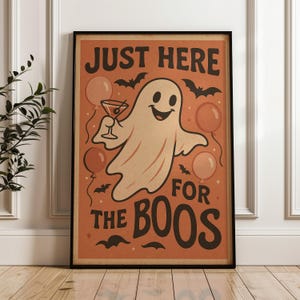 Op de afbeelding: Ingelijste Halloween-poster met een vintage ontwerp. De poster toont een vriendelijke geest met een cocktail, omringd door ballonnen en vleermuizen. De tekst luidt "JUST HERE FOR THE BOOS" in een vetgedrukte letter, tegen een oranje achtergrond.