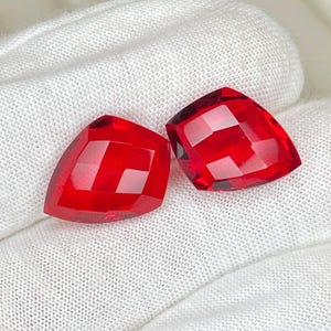Könnte beinhalten: Zwei facettierte, leuchtend rote Edelsteine mit dreieckiger Form. Die Edelsteine werden auf einem weißen, strukturierten Stoff präsentiert, der ihren brillanten Schliff und ihre Farbe zur Geltung bringt. Die Edelsteine sind wahrscheinlich für die Schmuckherstellung bestimmt.