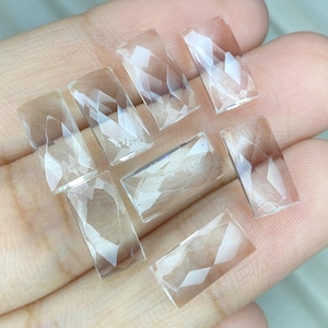 Pode incluir: Sete pedras preciosas transparentes, retangulares e facetadas dispostas em grupo. Cada pedra apresenta uma superfície superior plana com múltiplas facetas angulares ao longo das bordas, criando um padrão geométrico refletor de luz.