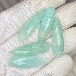 May include: Five faceted, pear-shaped amazonite gemstones in a pale mint green color. Each stone features a smooth, polished surface with subtle internal veining and a pointed tip, showcasing a translucent quality and a consistent light green hue.