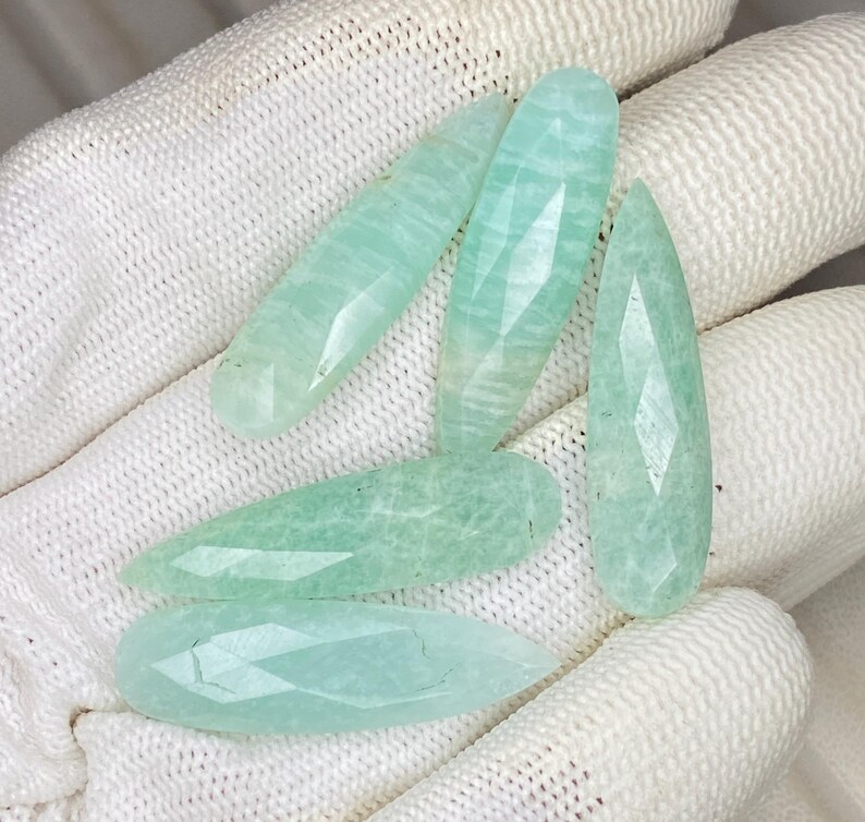 May include: Five faceted, pear-shaped amazonite gemstones in a pale mint green color. Each stone features a smooth, polished surface with subtle internal veining and a pointed tip, showcasing a translucent quality and a consistent light green hue.