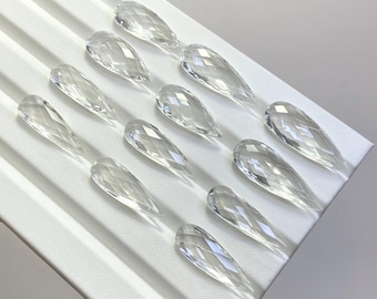 May include: Clear, faceted crystal teardrop-shaped pendants arranged on a white surface. The pendants are transparent, reflecting light and showcasing their intricate cuts. The arrangement highlights the pendants' shape and clarity.