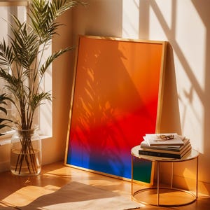 Könnte beinhalten: Ein rechteckiger gerahmter Druck mit einem lebendigen vertikalen Farbverlauf, der von einem warmen Orange oben über Rot zu einem tiefen Blau unten übergeht. Das Kunstwerk ist in einem dünnen goldenen Rahmen gefasst und steht auf dem Boden.