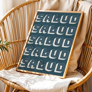 May include: A framed art print with the word "SALUD" repeated in a bold, white, 3D font against a teal background. The print is displayed on a woven chair with a neutral-colored cushion and a beige throw.