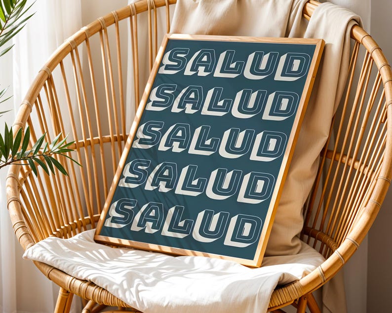 May include: A framed art print with the word "SALUD" repeated in a bold, white, 3D font against a teal background. The print is displayed on a woven chair with a neutral-colored cushion and a beige throw.