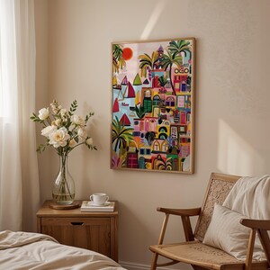 May include: A framed colorful artwork depicting a coastal town with sailboats, palm trees, and buildings. The artwork is in a wooden frame and hangs on a beige wall. A wooden chair, nightstand, and vase of flowers are in the room.