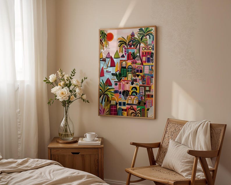 May include: A framed colorful artwork depicting a coastal town with sailboats, palm trees, and buildings. The artwork is in a wooden frame and hangs on a beige wall. A wooden chair, nightstand, and vase of flowers are in the room.