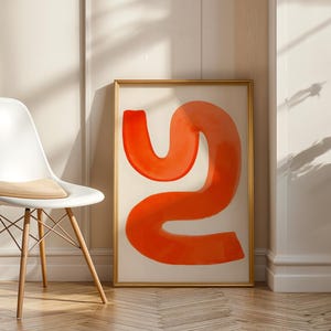 Könnte beinhalten: Ein abstrakter Kunstdruck mit einer leuchtend orangefarbenen, geschwungenen Form auf cremefarbenem Hintergrund, goldgerahmt. Links steht ein weißer Stuhl mit Holzbeinen und einem beigefarbenen Kissen. Das Kunstwerk lehnt an einer Wand mit Zierleisten.