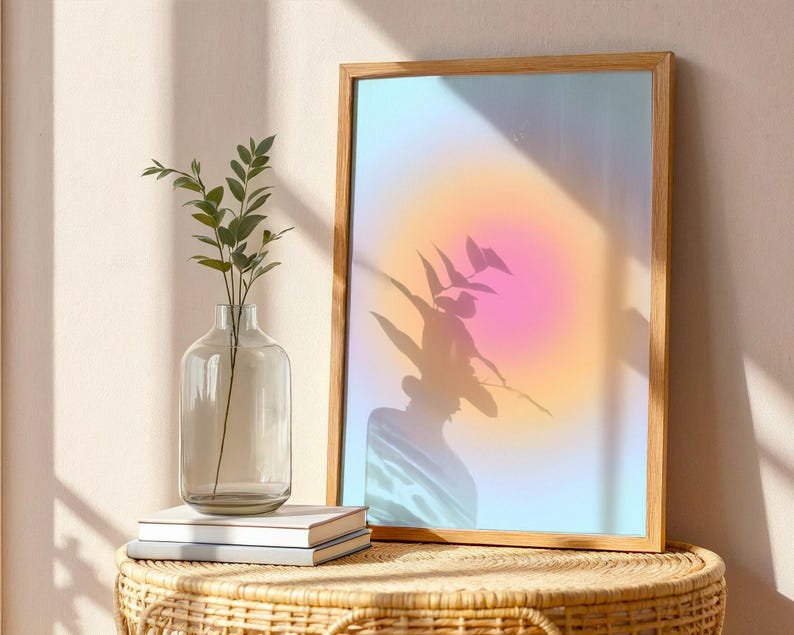 Puede incluir: Una impresi&oacute;n art&iacute;stica enmarcada con un dise&ntilde;o abstracto suave y colorido. La obra de arte presenta un degradado de tonos rosas, naranjas y azules. Un jarr&oacute;n de vidrio transparente con tallos y hojas verdes se encuentra sobre una mesa tejida, junto a una pila de libros.