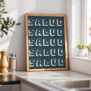 May include: A framed teal print with the word "SALUD" repeated in a 3D white font. The wooden frame sits on a countertop next to a salt shaker and a spice jar. The print is leaning against a white wall near a window.