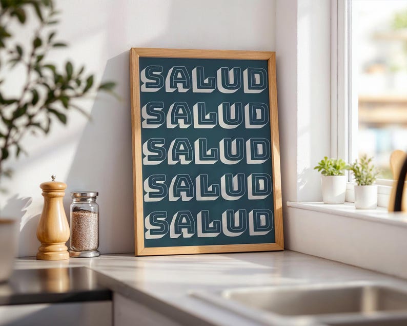 May include: A framed teal print with the word "SALUD" repeated in a 3D white font. The wooden frame sits on a countertop next to a salt shaker and a spice jar. The print is leaning against a white wall near a window.