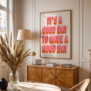 Könnte beinhalten: Ein gerahmter Kunstdruck mit dem Text "IT'S A GOOD DAY TO HAVE A GOOD DAY" in rosa und orangefarbenen Blockbuchstaben. Der Druck hängt über einem Holzschrank mit Büchern und einer Lampe.