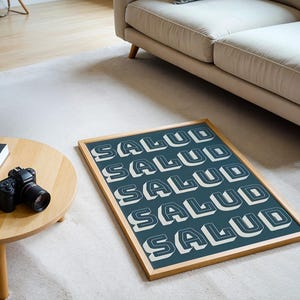 May include: A framed art print with the word "SALUD" in white, 3D block letters repeated across a teal background. The wooden frame sits on a white rug near a wooden table with a camera and books.