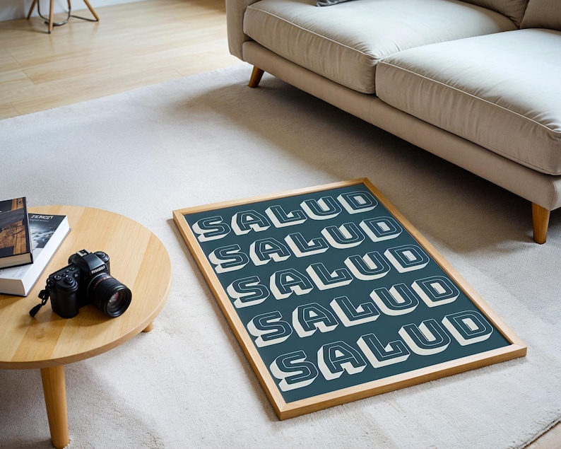 May include: A framed art print with the word "SALUD" in white, 3D block letters repeated across a teal background. The wooden frame sits on a white rug near a wooden table with a camera and books.