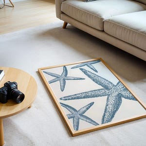 Könnte beinhalten: Ein gerahmter Kunstdruck mit mehreren blauen Seesternen auf cremefarbenem Hintergrund. Das Kunstwerk wird auf einem weißen Teppich in der Nähe eines hölzernen Couchtisches und eines beigen Sofas ausgestellt. Der Rahmen besteht aus hellem Holz.