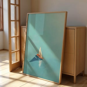 Könnte beinhalten: Ein gerahmter Kunstdruck mit einem Segelboot auf türkisblauem Hintergrund. Das Segelboot hat einen orangefarbenen Rumpf und ein weißes Segel. Der Rahmen ist helles Holz. Das Kunstwerk lehnt an einem hellen Holzschrank.