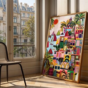 May include: A colorful framed art print depicting a vibrant coastal town scene with boats, palm trees, and buildings in shades of pink, yellow, blue, and green. The artwork is leaning against a wall next to a window with a view of a city.