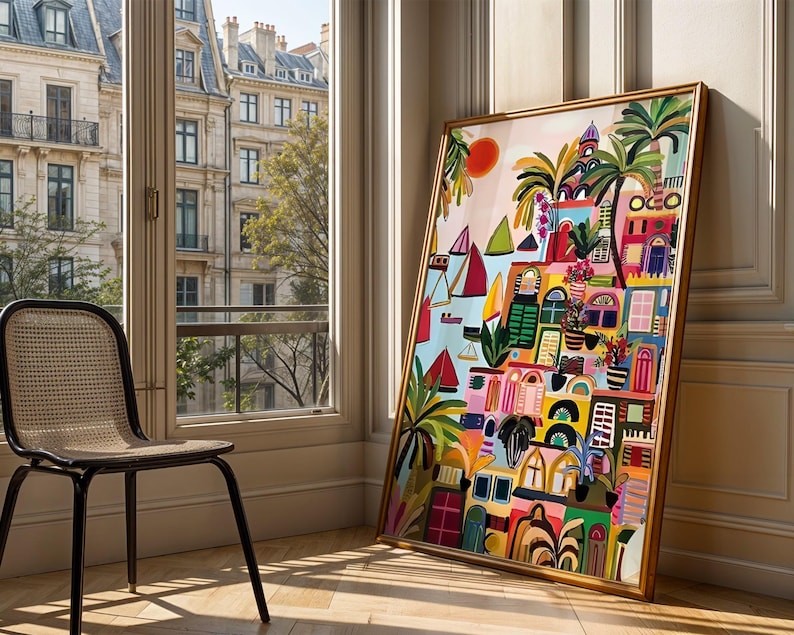 May include: A colorful framed art print depicting a vibrant coastal town scene with boats, palm trees, and buildings in shades of pink, yellow, blue, and green. The artwork is leaning against a wall next to a window with a view of a city.