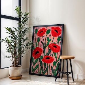 Könnte beinhalten: Ein gerahmter Kunstdruck mit einem floralen Design aus roten Mohnblumen auf rosa Hintergrund. Das Kunstwerk wird vor einer weißen Wand präsentiert, neben einer Topfpflanze und einem Holzschemel mit schwarzen Beinen. Die Szene hat eine moderne, dekorative Ausstrahlung.